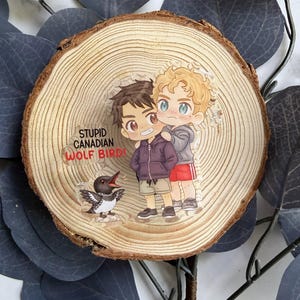 May include: A clear pin featuring two cartoon figures embracing, with a cartoon loon bird. The pin is on a wooden slice. Text on the pin reads "STUPID CANADIAN WOLF BIRD!"