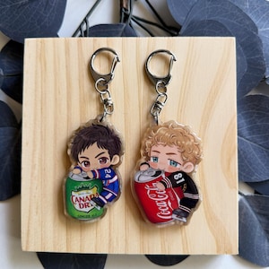 May include: Two acrylic keychains featuring cartoon characters. One keychain depicts a character with brown hair holding a Canada Dry Ginger Ale can. The other shows a character with blonde hair holding a Coca-Cola can. Both keychains have silver key rings.