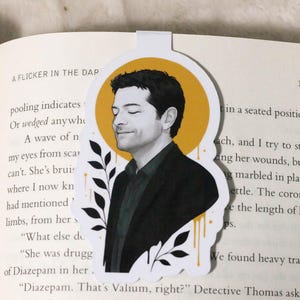 May include: Bookmark featuring a black and white illustration of a man with a yellow halo and black leaves. The bookmark is placed on a book with text visible.