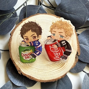 Könnte beinhalten: Zwei transparente Acryl-Charms mit Cartoon-Figuren, die jeweils eine Limonadendose halten. Eine Figur hält eine grüne "Canada Dry Ginger Ale" Dose, die andere eine rote "Coca-Cola" Dose. Die Charms liegen auf einer Holzscheibe.