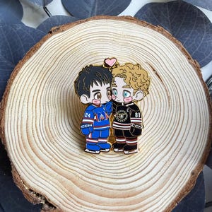 May include: A gold-colored enamel pin featuring two cartoon hockey players embracing, with a pink heart above them. One player wears a blue jersey with the number 24, the other a black and red jersey with the number 81. The pin is set on a wooden slice.