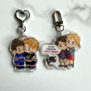 May include: Two acrylic keychains featuring cartoon illustrations of two people. One keychain shows two figures in hockey jerseys, the other shows two figures with the text "STUPID CANADIAN WOLF BIRD!" and a bird graphic. Both have metal hardware.