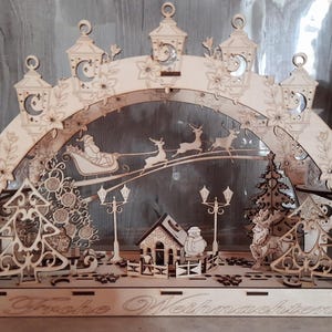 Puede incluir: Decoración navideña de madera con un diseño detallado cortado con láser. La escena incluye a Papá Noel en un trineo tirado por renos, una casa, árboles y farolas. El arco está adornado con faroles y detalles florales. Las palabras "Frohe Weihnachten" están en la base.