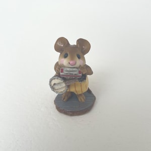 May include: A small brown mouse figurine standing on a gray circular base. The mouse has large ears, a pink nose, and rosy cheeks. It is wearing a yellow striped outfit and is holding a silver harmonica to its mouth while playing a small banjo.
