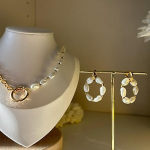 Puede incluir: Un collar de cadena dorada con un gran cierre de anillo dorado y una hebra de cuentas blancas de forma ovalada. Pendientes de aro dorados a juego con cuentas ovaladas blancas se exhiben en un soporte dorado. La joyería se presenta sobre un busto blanco y un soporte dorado.