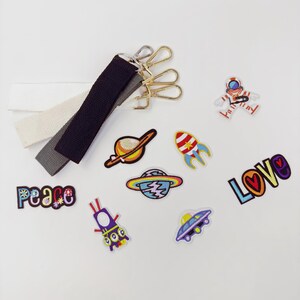 May include: An assortment of colorful embroidered patches and three canvas straps with gold-tone metal clasps. Patches include a rocket, planet, astronaut, and the words "peace" and "love". The straps are white, gray, and black.
