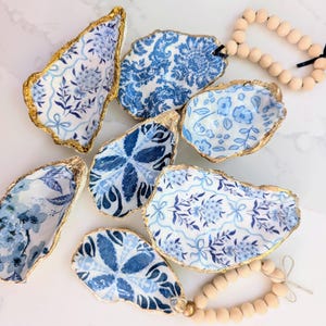 May include: A collection of decorative oyster shell dishes with blue floral patterns and gold-painted edges. The shells vary in size and shape, and are arranged on a white surface. A wooden bead garland with a black ribbon is also present.