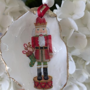 May include: A Christmas ornament featuring a nutcracker design on a white, shell-shaped base with gold trim. The nutcracker is dressed in a red coat and black boots, holding a stocking. A red ribbon and white bead are attached for hanging.