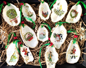 May include: A set of twelve oyster shell ornaments, each featuring a unique hand-painted illustration representing the Twelve Days of Christmas, numbered one through twelve, with gold-painted edges and green ribbons with red beads for hanging.