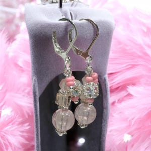 May include: A pair of dangle earrings with silver-tone lever-back closures. Each earring features a cluster of pink and clear beads, including faceted crystals and round, light pink beads. The earrings are displayed against a pink feathered background.