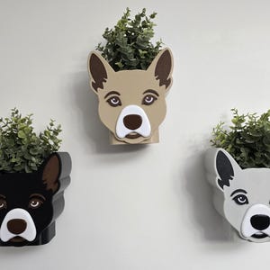 May include: Three dog-shaped wall planters, each with a different color scheme. One is black, one is tan, and one is gray. Each planter holds a cluster of green faux foliage. The planters are mounted on a white wall.