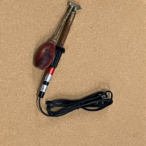 May include: A soldering iron with a wooden handle and a metal tip. The iron has a black power cord and is set against a neutral background.