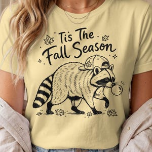 Könnte beinhalten: Ein hellgelbes T-Shirt mit einer schwarzen Strichzeichnung eines Waschbären, der eine Baseballkappe trägt und eine Blase bläst. Der Text "Tis The Fall Season" ist darüber gedruckt, umgeben von kleinen Ahornblatt- und Sternakzenten.