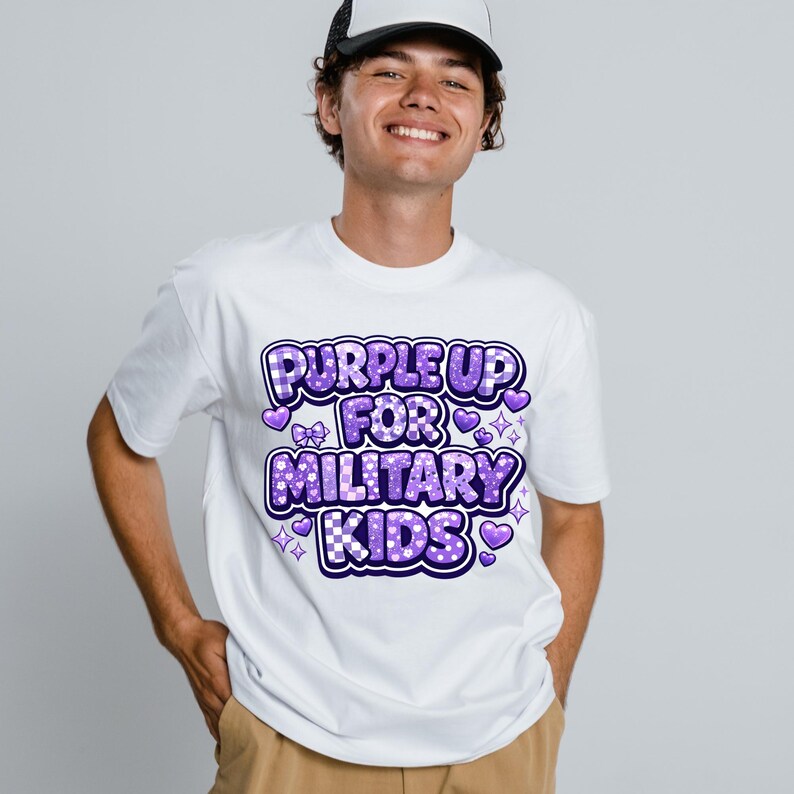 May include: White t-shirt with the text "PURPLE UP FOR MILITARY KIDS" in purple, featuring hearts, bows, and stars. The shirt is short-sleeved and has a crew neck.