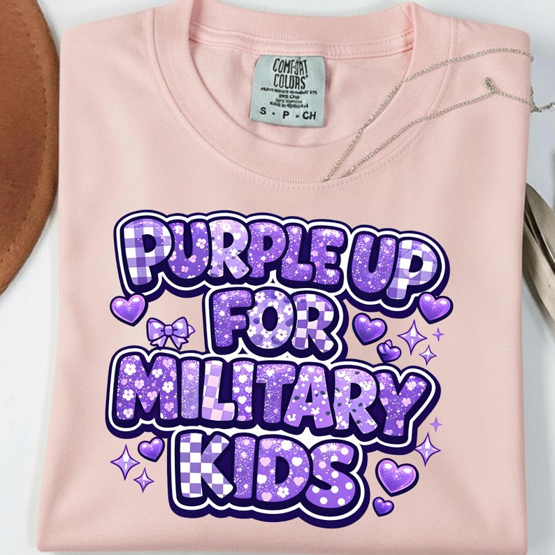 May include: A light pink t-shirt with the text "PURPLE UP FOR MILITARY KIDS" in purple, white, and blue. The design includes hearts, stars, and a bow. The shirt has a Comfort Colors tag.