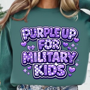 May include: Teal sweatshirt with the text "PURPLE UP FOR MILITARY KIDS" in a purple, checkered, and floral design. The design includes hearts, bows, and stars. The sweatshirt is a solid teal color.