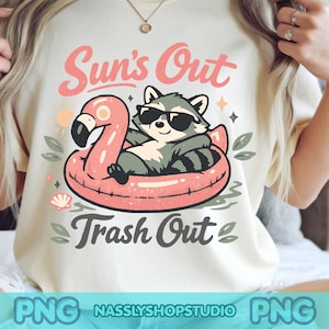 May include: A cream-colored t-shirt featuring a graphic of a raccoon wearing black sunglasses while floating in a pink flamingo pool float. The text above reads Sun's Out and the text below reads Trash Out in a dark gray script font.