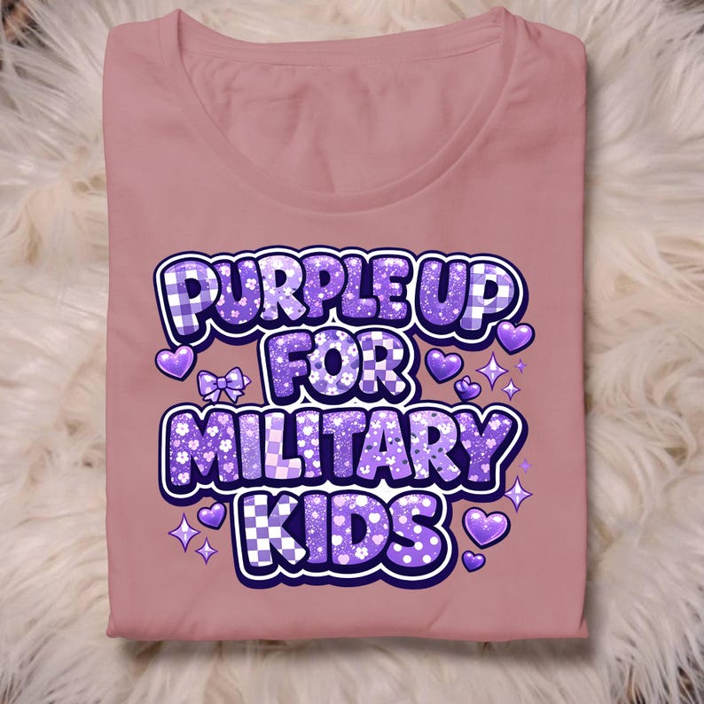 May include: A mauve-colored t-shirt with the text "PURPLE UP FOR MILITARY KIDS" in a purple, white, and blue design. The design includes hearts, stars, and a bow. The shirt is folded.