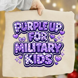 May include: A beige pillow with the text "PURPLE UP FOR MILITARY KIDS" in purple. The text has a mix of patterns, including plaid and floral designs, with purple hearts, bows, and stars. The pillow is held by a person.