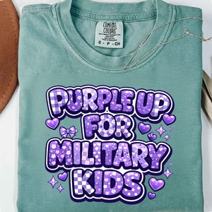 May include: A seafoam green t-shirt with the text "PURPLE UP FOR MILITARY KIDS" in purple, white, and blue. The design includes hearts, bows, and stars. The shirt has a crew neck and short sleeves.