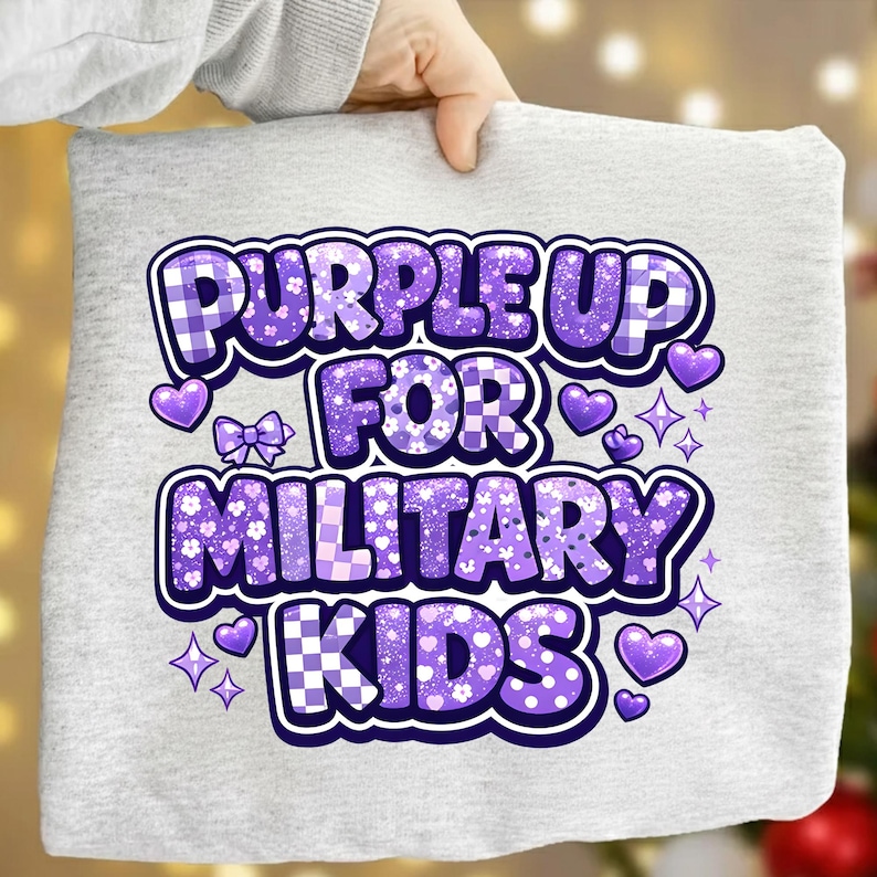 May include: A light gray t-shirt with the text "PURPLE UP FOR MILITARY KIDS" in a purple, decorative font. The text is surrounded by purple hearts, stars, and bows. The shirt is held by a person.