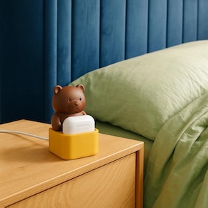 May include: A brown bear-shaped charging station with a yellow base holds a white charging case. The station sits on a wooden nightstand next to a bed with a green pillow and blue headboard.