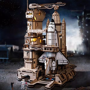 May include: A detailed wooden model of a space shuttle and launch tower, with intricate cutouts and a light-up interior. The model features a white and black shuttle, a launch pad, and a tower with multiple levels. The scene is set against a backdrop of a starry night.
