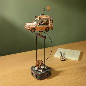 Puede incluir: Maqueta de madera de un vehículo todoterreno naranja y blanco, con la inscripción "Sunny Day". El modelo está suspendido sobre una base con un diseño de globo terráqueo y una rueda giratoria. También hay un pequeño ukelele y un libro.