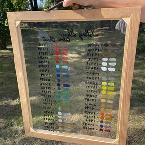 May include: A wooden-framed color chart with a clear glass panel. The chart displays various paint color swatches in vertical columns. The colors range from reds and blues to greens and yellows. Handwritten text is visible on the glass.