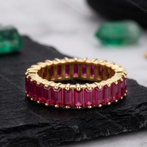 May include: A gold eternity band featuring a continuous row of vibrant, rectangular-cut magenta rubies set in a shared-prong metal setting. The ring displays a uniform, repeating pattern of gemstones around the entire circumference of the band.