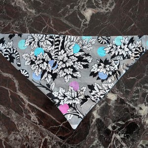 May include: A triangular pet bandana with a black plastic buckle. The bandana features a gray background with a floral and animal print in black and white, accented with blue, pink, and purple polka dots. The design includes stylized flowers and lemurs.