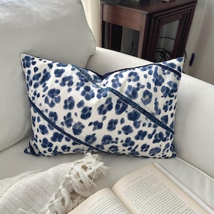 May include: A decorative pillow with a white background and a blue leopard print pattern. The pillow has a dark blue border and diagonal stripes. The pillow is on a white couch.