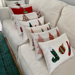 K&ouml;nnte beinhalten: Eine Sammlung von Weihnachtskissen mit verschiedenen Designs, darunter Weihnachtsb&auml;ume, Rentiere und das Wort "JOY". Die Kissen sind haupts&auml;chlich wei&szlig; mit roten, gr&uuml;nen und braunen Akzenten, angeordnet auf einem wei&szlig;en Sofa.