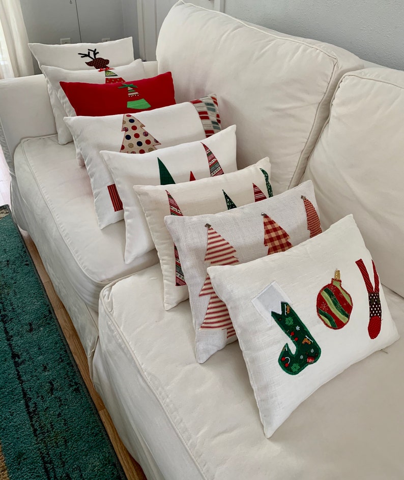 K&ouml;nnte beinhalten: Eine Sammlung von Weihnachtskissen mit verschiedenen Designs, darunter Weihnachtsb&auml;ume, Rentiere und das Wort "JOY". Die Kissen sind haupts&auml;chlich wei&szlig; mit roten, gr&uuml;nen und braunen Akzenten, angeordnet auf einem wei&szlig;en Sofa.