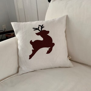 K&ouml;nnte beinhalten: Quadratisches wei&szlig;es Kissen mit einem braunen Rentier-Design. Das Rentier besteht aus gemustertem Stoff. Das Kissen liegt auf einem wei&szlig;en Sofa. Ein dekorativer Artikel f&uuml;r das Zuhause.