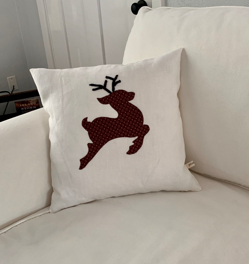 K&ouml;nnte beinhalten: Quadratisches wei&szlig;es Kissen mit einem braunen Rentier-Design. Das Rentier besteht aus gemustertem Stoff. Das Kissen liegt auf einem wei&szlig;en Sofa. Ein dekorativer Artikel f&uuml;r das Zuhause.