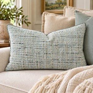 May include: A rectangular throw pillow featuring a textured, woven fabric in shades of light blue, cream, and dark navy. The pillow has a dense, multi-colored tweed-like pattern with visible horizontal threads and a clean, piped edge finish.
