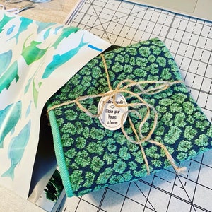 K&ouml;nnte beinhalten: Ein Stoffb&uuml;ndel mit gr&uuml;nem und marinefarbenem Leopardenmuster, mit Bindfaden gebunden und einem Etikett mit der Aufschrift "Make your house a home". Das B&uuml;ndel befindet sich neben einem Blatt Papier mit Aquarellfisch-Illustrationen. Der Stoff hat einen gr&uuml;nen Rand.