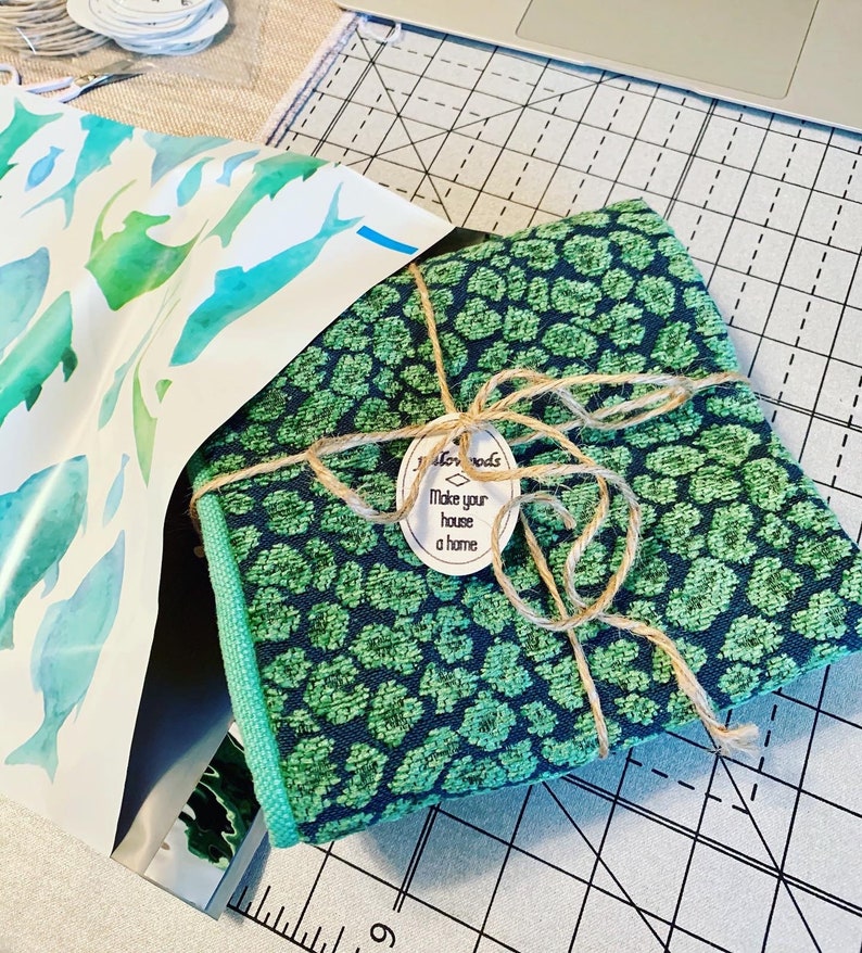 K&ouml;nnte beinhalten: Ein Stoffb&uuml;ndel mit gr&uuml;nem und marinefarbenem Leopardenmuster, mit Bindfaden gebunden und einem Etikett mit der Aufschrift "Make your house a home". Das B&uuml;ndel befindet sich neben einem Blatt Papier mit Aquarellfisch-Illustrationen. Der Stoff hat einen gr&uuml;nen Rand.
