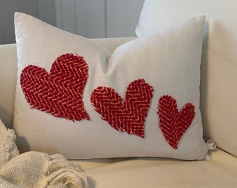 May include: White rectangular pillow with three red heart appliqués. The hearts have a textured, herringbone pattern. The pillow is on a white couch, with a white throw blanket visible in the foreground.