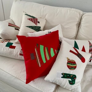K&ouml;nnte beinhalten: Eine Sammlung von Weihnachtskissen in verschiedenen Formen und Gr&ouml;&szlig;en. Die Kissen zeigen Applikationsdesigns von Weihnachtsb&auml;umen, Ornamenten und abstrakten Mustern in Rot, Gr&uuml;n und Wei&szlig; auf wei&szlig;em Hintergrund. Die Kissen sind auf einem wei&szlig;en Sofa angeordnet.