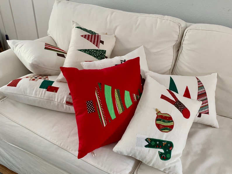K&ouml;nnte beinhalten: Eine Sammlung von Weihnachtskissen in verschiedenen Formen und Gr&ouml;&szlig;en. Die Kissen zeigen Applikationsdesigns von Weihnachtsb&auml;umen, Ornamenten und abstrakten Mustern in Rot, Gr&uuml;n und Wei&szlig; auf wei&szlig;em Hintergrund. Die Kissen sind auf einem wei&szlig;en Sofa angeordnet.