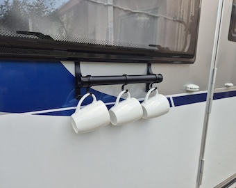 Könnte beinhalten: Ein schwarzer Metall-Becherhalter mit drei weißen Keramikbechern, die daran hängen. Der Halter ist an der Seite eines Fahrzeugs montiert, mit einem blau-weißen Streifendesign. Die Becher haben einen einfachen, gebogenen Griff.