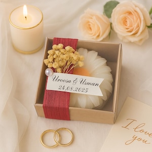 Peut inclure: Une boîte de faveur de mariage avec une bougie en forme de marguerite, des fleurs séchées et une étiquette indiquant "Uroosa & Usman 24.08.2025". Des alliances dorées et une bougie allumée sont également présentes. Des roses pêche et une carte "I love you" en arrière-plan.