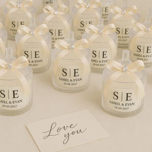 Op de afbeelding: Glazen kaarsjargen met cr&egrave;mekleurige strikken en kristallen accenten. Elke pot heeft de initialen "SE", de namen "Sahel & Evan" en een datum. Een kaart met "Love you" ligt op de voorgrond.