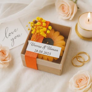 Peut inclure: Une bougie en forme de tournesol dans une boîte transparente, ornée de fleurs jaunes et d'un ruban orange. La boîte porte une étiquette avec les noms "Uroosa & Usman" et la date "24.08.2025". Des alliances dorées et une bougie allumée sont également visibles.