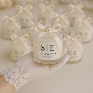 Peut inclure: Plusieurs petits pots à bougies en verre transparent avec des bougies crème à l'intérieur. Chaque pot est orné d'un ruban crème et d'un petit bijou. L'avant de chaque pot porte les initiales "S E", les noms "SAHEL & EVAN" et la date "29.09.2027".