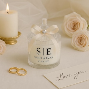 Op de afbeelding: Een brandende kaars en een decoratieve glazen pot met een cr&egrave;mekleurig lint en een kristallen accent. De pot heeft de initialen "S|E" en de namen "SAHEL & EVAN" met de datum "29.09.2027" erop gedrukt. Twee gouden trouwringen en een "Love you"-kaart zijn ook aanwezig.