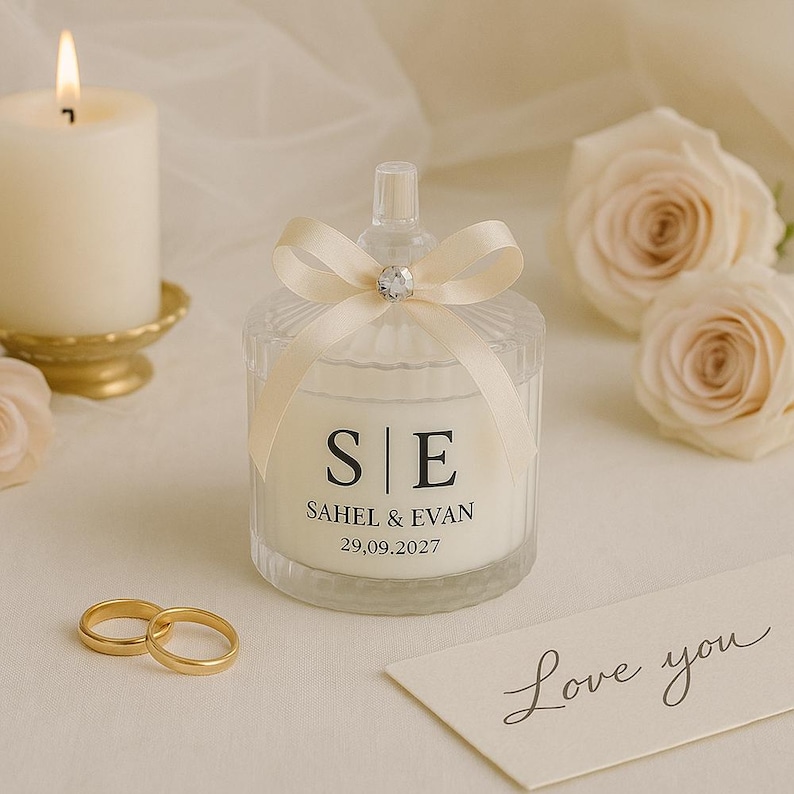 Op de afbeelding: Een brandende kaars en een decoratieve glazen pot met een cr&egrave;mekleurig lint en een kristallen accent. De pot heeft de initialen "S|E" en de namen "SAHEL & EVAN" met de datum "29.09.2027" erop gedrukt. Twee gouden trouwringen en een "Love you"-kaart zijn ook aanwezig.