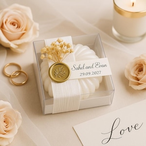 Può includere: Una candela bianca e rotonda in una scatola trasparente, decorata con un sigillo dorato e fiori secchi. Il sigillo ha un disegno a foglie e il testo "Sahel and Evan 29.09.2027". È un segnaposto per matrimoni.